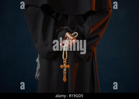 Male monk in black robe holds wooden rosary and cross in hands, religion. Mysterious friar in dark cape Stock Photo