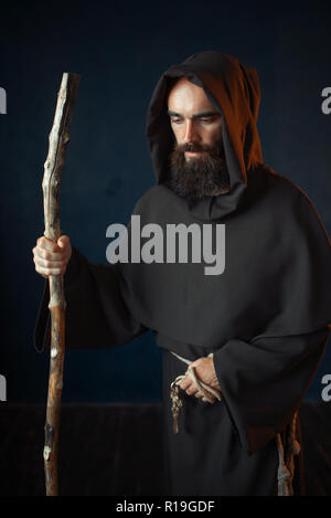 Medieval monk in black robe with hood rests on a stick, religion. Mysterious friar in dark cape Stock Photo