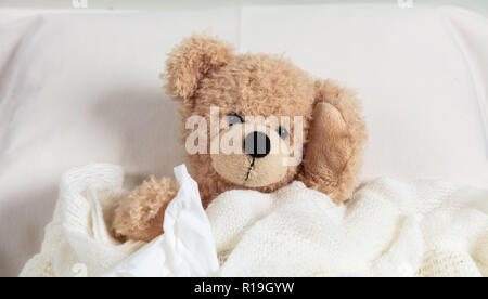 Cold, flue or allergy concept. Cute teddy in bed, covered with a warm blanket, holding a tissue Stock Photo