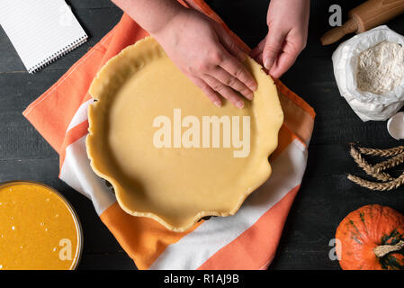 Woman puts the pie crust in a baking tray, on a kitchen towel, and pumpkin pie filling, flour, rolling pin, on a rustic table. Holiday home baking. Stock Photo
