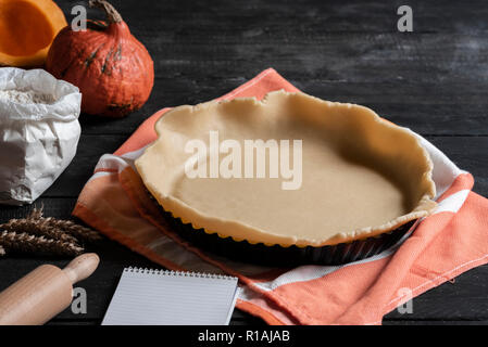 Holiday dessert preparation with the pie crust in a baking tray, on a towel, with ingredients like pumpkin, flour, and an empty notebook. Stock Photo