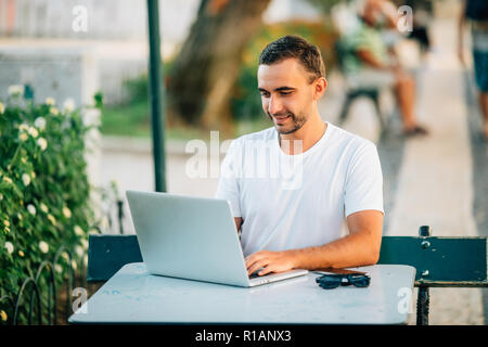 Young handsome male using a laptop in a cafe on a street. Stock Photo