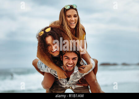 Man giving piggyback ride to female friends on beach. Happy young friends having fun on the seashore, enjoying summer holidays. Stock Photo