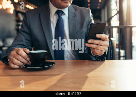Cropped shot of smart phone in hand of a senior businessman with coffee on cafe table. Senior businessman texting on mobile phone at coffee shop. Stock Photo