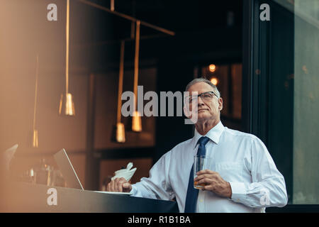 Thoughtful senior man with glass of beer in hand sitting at cafe counter looking away. Mature male entrepreneur looking away thinking while having a b Stock Photo