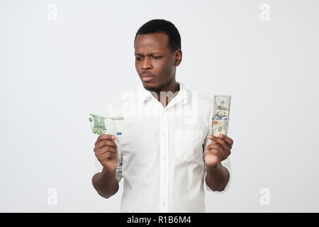 Young attractive african man holding USA currency and euro currency deciding which one to choose. How is it better to save money concept Stock Photo