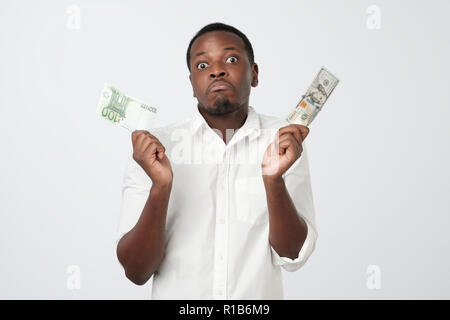 Young attractive african man holding USA currency and euro currency deciding which one to choose. How is it better to save money concept Stock Photo