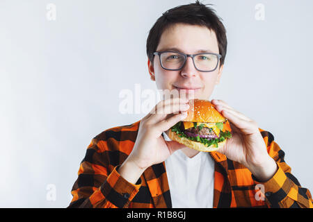 A young guy with glasses holding a fresh Burger. A very hungry student eats fast food. Hot helpful food. The concept of gluttony and unhealthy diet. With copy space for text Stock Photo
