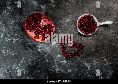 Tempting pomegranate, red seeds arranged in the shape of a heart and fruit on a black, stone background Stock Photo