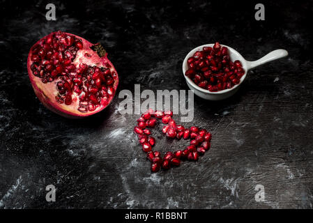 Tempting pomegranate, red seeds arranged in the shape of a heart and fruit on a black, stone background Stock Photo