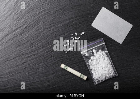 white crystals of drugs in a bag on a black table. next to it is a $ 100 bill folded into a tube Stock Photo