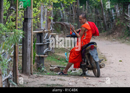 Don Det, Laos - April 23, 2018: Young buddhist monk sitting on a scooter and using his smartphone in a remote village of southern Laos Stock Photo