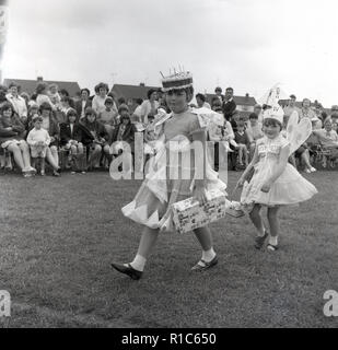 1967, historcial, outside in a fieldd, at an English village fete, two young girls dressed in costumes walk infront of the spectators in a fancy dress competition, England, UK. One of the girls has a party dress on and wearing a cake on her head, while the other is dressed as an angel and has the words Gipsy Moth IV on her, the name of the boat Sir Francis Chichester who the following month, successfuly sailed, single-handed, around the world. Her thoughts were with him. Stock Photo