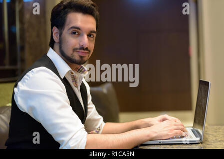 Young handsome Indian man using laptop in the lobby of hotel Stock Photo