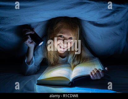 Happy smart girl reading a fairytale book lying in the dark under the cover bed holding a lantern in Reading skills Literature School Success and Educ Stock Photo