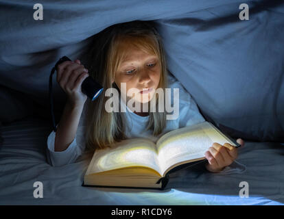 Beautiful caucasian girl lying in bed under the duvet holding a lantern reading a mystery book in the dark late at night looking frightened with a dra Stock Photo