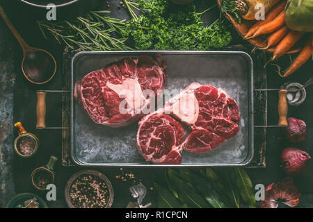 Raw beef meat shin with bone for broth, soup or stew in metal tray with ingredients for tasty cooking: root vegetables, herbs and spices on dark rusti Stock Photo