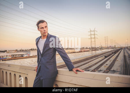 A handsome male model dressed in a grey suit standing by a bridge in Los Angeles at dusk with a beautiful skyline. He looks away and looks ambitious. Stock Photo