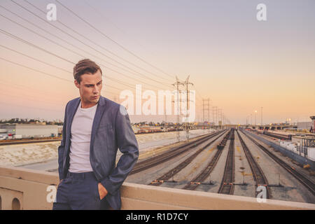 A handsome male model dressed in a grey suit standing by a bridge in Los Angeles at dusk with a beautiful skyline. He looks down, deep in thought. Stock Photo