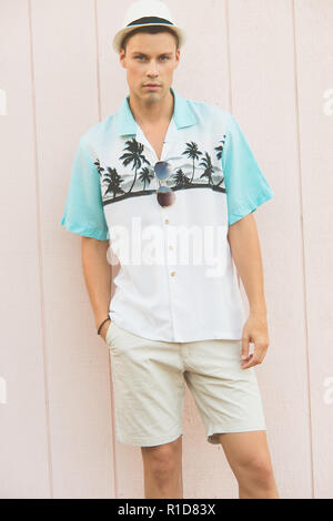A handsome male model posing in Summer Clothes outdoor against a plain wooden wall. He wears a white fedora hat, looking confident. Stock Photo