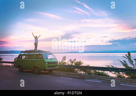 Young beautiful girl standing on the roof of old timer classic camper van parked by the sea with amazingly colorful sunset sky, hands wide open free Stock Photo