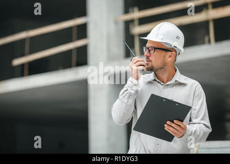 Male head engineer wearing white safety hardhat with walkie talkie and clipboard inspecting construction site. Development and construction industry c Stock Photo