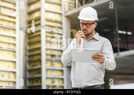 Male head engineer wearing white safety hardhat with walkie talkie and tablet inspecting construction site. Technology, development and construction i Stock Photo