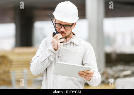 Male head engineer wearing white safety hardhat with walkie talkie and tablet inspecting construction site. Technology, development and construction i Stock Photo
