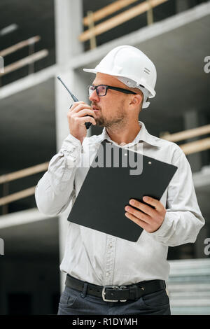 Male head engineer wearing white safety hardhat with walkie talkie and clipboard inspecting construction site. Development and construction industry c Stock Photo