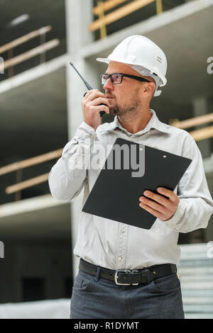 Male head engineer wearing white safety hardhat with walkie talkie and clipboard inspecting construction site. Development and construction industry c Stock Photo