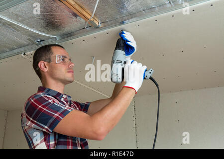 Young man in goggles fixing drywall suspended ceiling to metal frame using electrical screwdriver on ceiling insulated with shiny aluminum foil. Renov Stock Photo