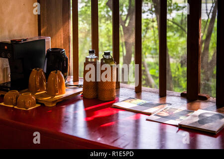 A coffee machine, tea cups and two rattan-wrapped water bottles stand on a red counter by the slatted window of a hotel room at the eco-lodge Jetwing Kaduruketha near Wellawaya, Sri Lanka, with magazines open in front of them. Stock Photo