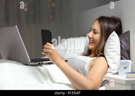 Happy woman using a smart phone and laptop lying on a bed in the night at home Stock Photo
