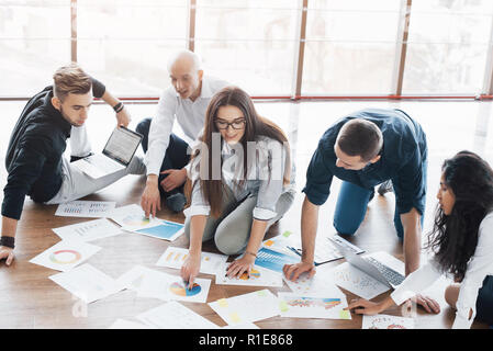 Planning strategy together. Business team looking at papers on floor with manager pointing to one idea. Cooperation corporate achievement. Planning design draw. Teamwork concept Stock Photo