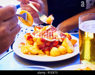 People taste a portion of Patatas Bravas over a metallic table. Fried potatoes topped with spicy sauce, one of the most common typical Spanish tapas. Stock Photo