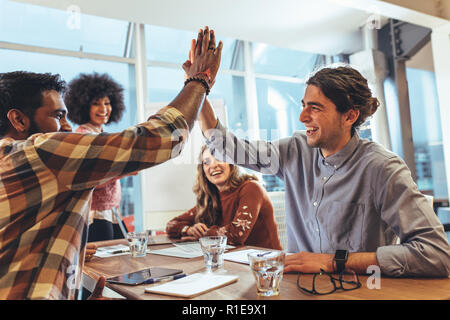 Coworkers sitting in conference room celebrating success. businessman giving a high five to his coworker while their female colleagues look on. Stock Photo