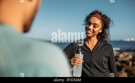 Smiling woman relaxing after a workout drinking water. Woman talking to her friend after workout standing outdoors. Stock Photo