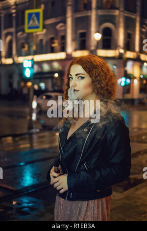 Young red-haired girl smokes an electronic cigarette. She is walking in street of night city. Wet asphalt after rain. Stock Photo