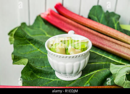 Rhubarb (Rheum rhabarbarum) peeled sliced and cut in to pieces in a white cup on a rhubarb leaf and with rhubarb stalks. White wooden background. Stock Photo