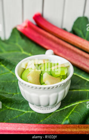 Rhubarb (Rheum rhabarbarum) peeled sliced and cut in to pieces in a white cup on a rhubarb leaf and with rhubarb stalks. White wooden background. Stock Photo