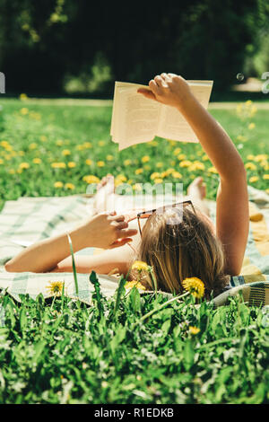 Young woman lying down on a blanket in home yard and sunbathing on a sunny day and reading a real book. Grass and dandilion background, nice vintage. Stock Photo