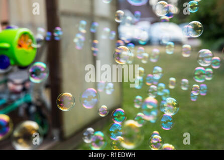 A lot of colorful small soap bubbles fly out of the bubble machine toy outdoors, green trees background. Stock Photo