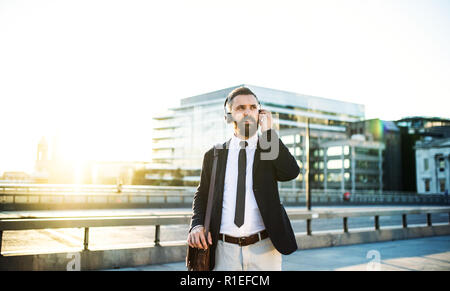 Hipster businessman with hadphones standing outdoors in the city at sunset. Stock Photo