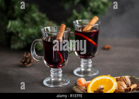 Winter composition. Mulled wine in glasses at black background. Fir wreath, tray with orange, cinnamon, nuts, cone and spices near. Stock Photo
