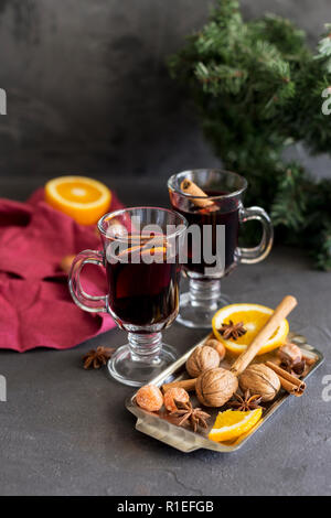 Winter christmas composition. Red mulled wine in glasses at black background. Fir wreath, tray with orange, cinnamon, nuts, cone and spices near.