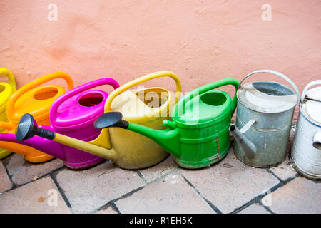 Old and used plastic and metal watering cans and pots in various colors for watering plants and flowers at home and garden stand on a stone tiles near Stock Photo
