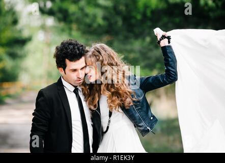 Cute couple embraces. Portrait of stylish young man in suit with tie hugs beautiful girl holding white dress in air. Concept of successful relationshi Stock Photo