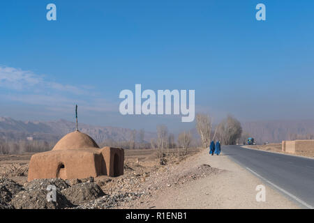 2 Ladies Wearing Blue Burqa Walking On Road Entering The Bamyan Valley With A Shrine And Mountains In The Background, Bamyan Province, Afghanistan Stock Photo