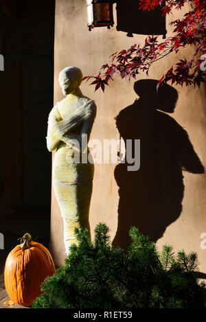 A spooky authentic looking gauze wrapped Mummy stands in front of an adobe house at sunset along with a big pumpkin for Halloween fun Stock Photo