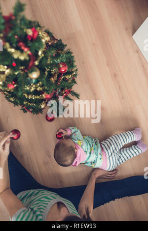 Top view of a mother sitting on living room floor decorating Christmas tree with her baby daughter lying next to her playing with red holiday bauble.  Stock Photo
